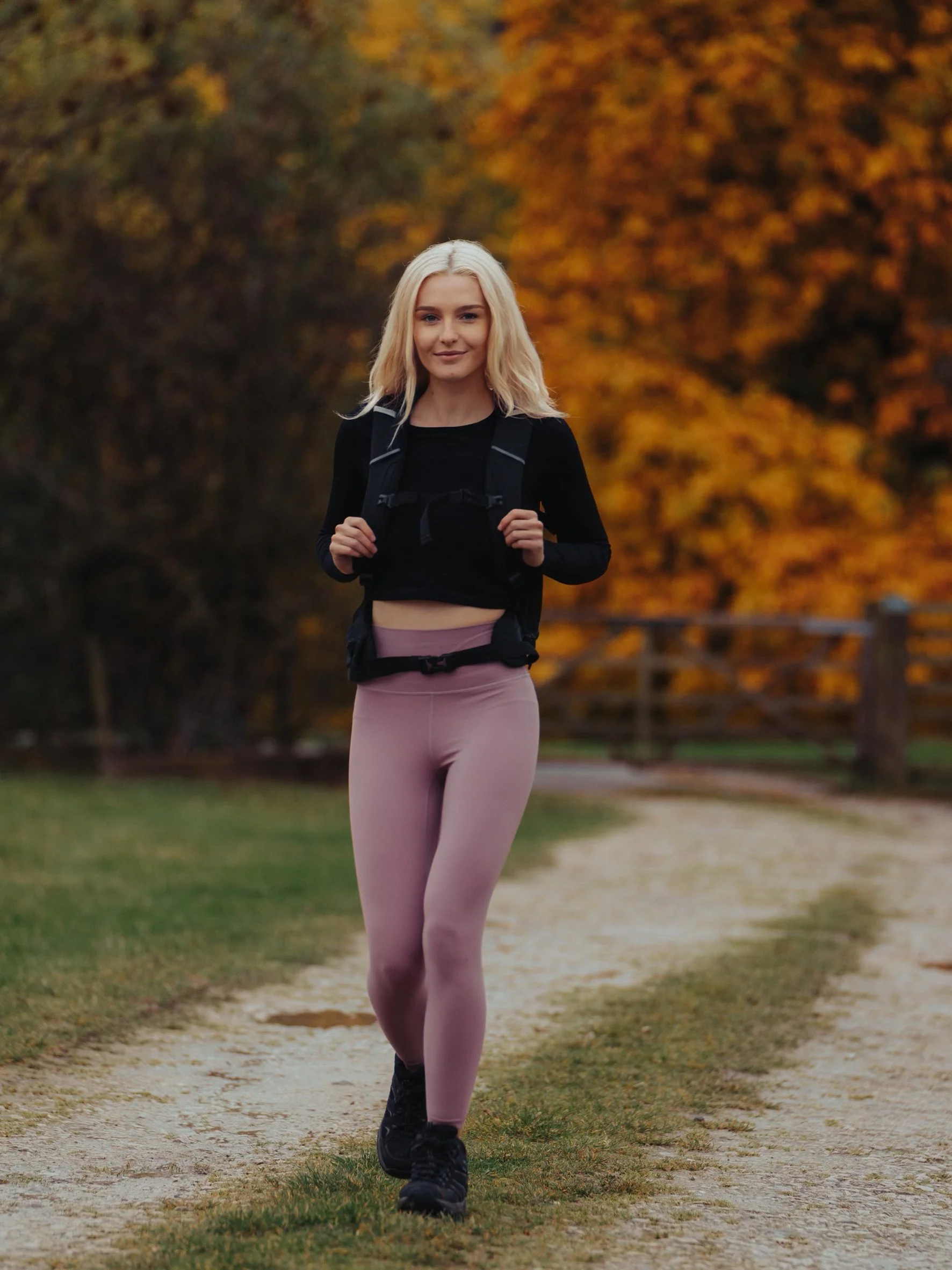A young woman with blonde hair, wearing a black top and pink leggings, jogging on a dirt path in a park during autumn with yellow and orange leaves in the background.