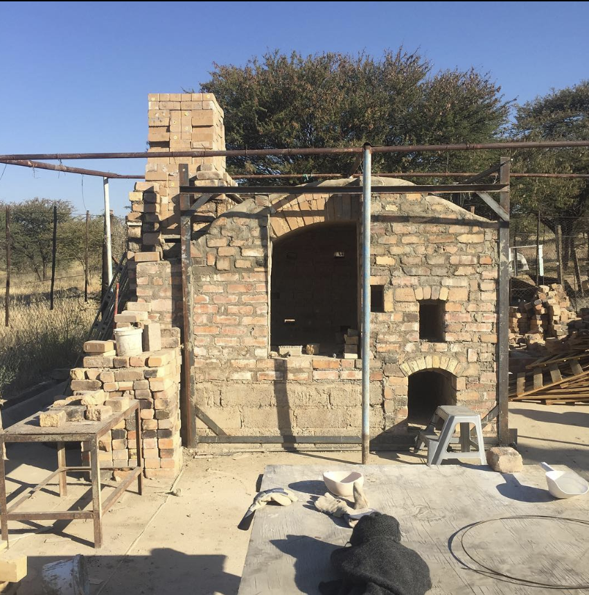 Construction site with a brick kiln in progress, surrounded by tools and materials, with trees and blue sky in the background.