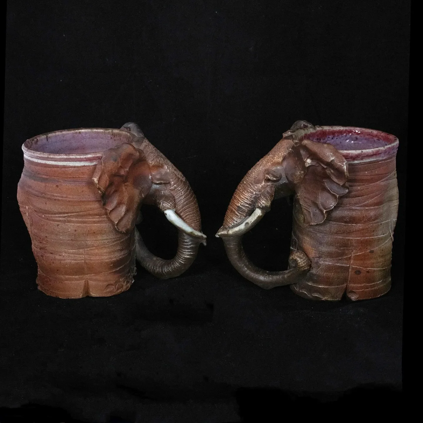 Two ceramic mugs with elephant head designs, featuring sculpted elephant ears and trunks, against a black background.