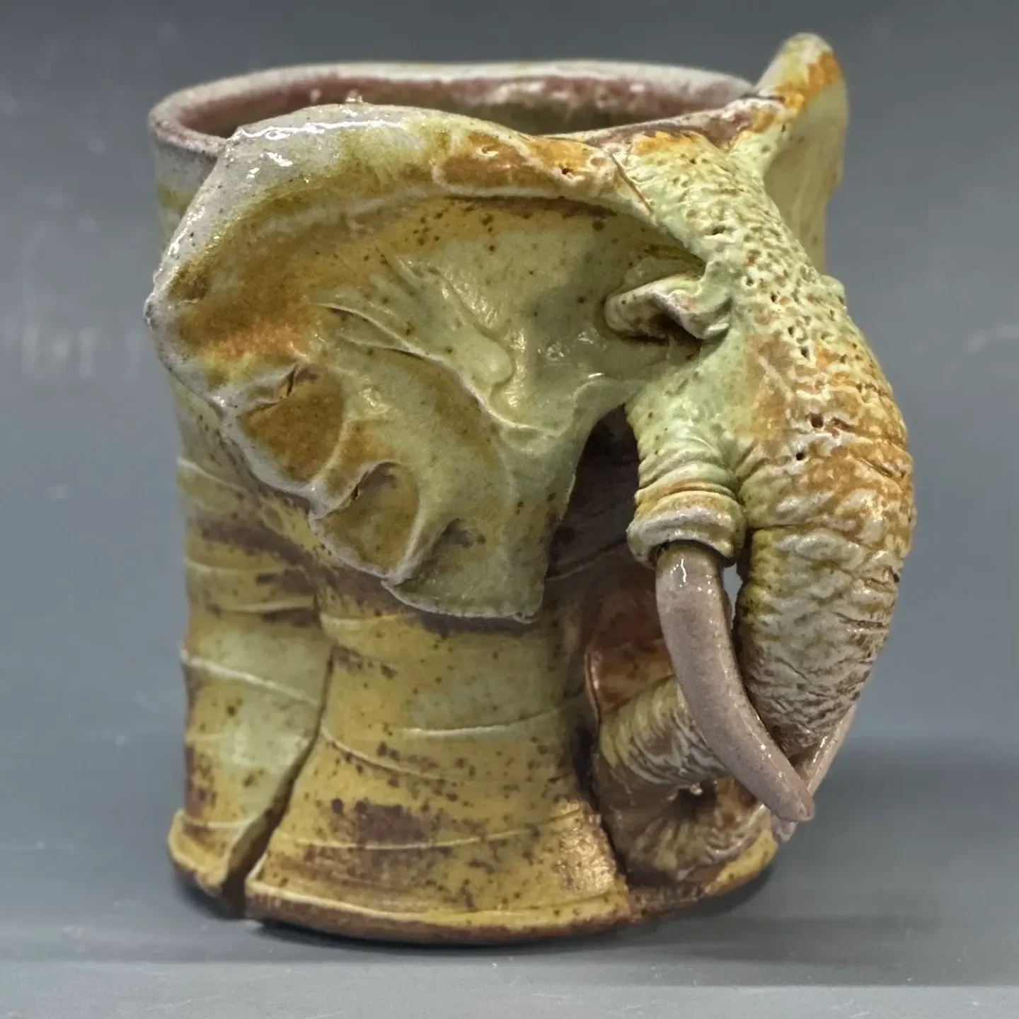A ceramic mug shaped like an old, weathered elephant with a small, curved trunk and textured details, displayed against a plain background.