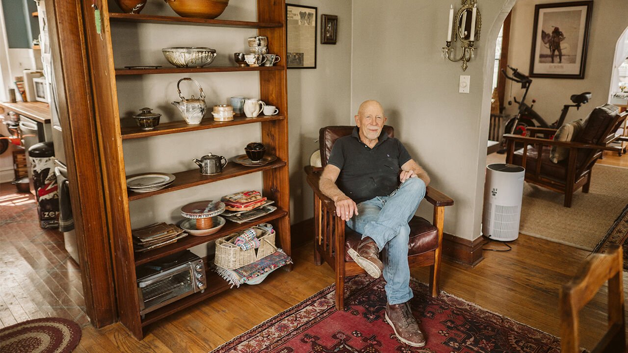 sitting comfortably in a wooden armchair in a cozy living room with hardwood flooring and a patterned rug. Behind him is a wooden bookshelf filled with decorative bowls, plates, and other dishware. To the right, there is a white air purifier, and in the background, there is a workout bike in a separate room.