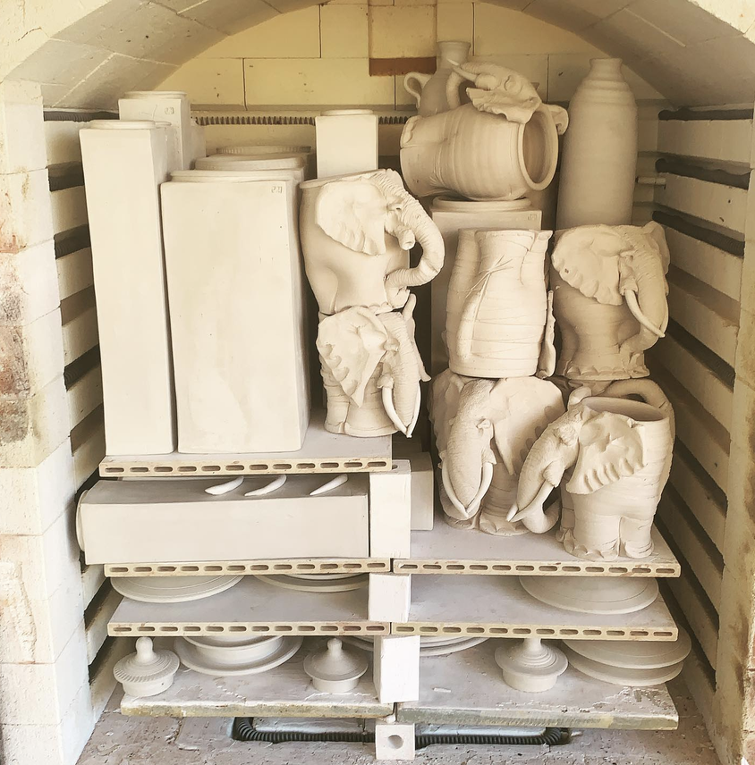 Unfinished ceramic sculptures of elephants, vases, and other pottery items on shelves in a kiln.