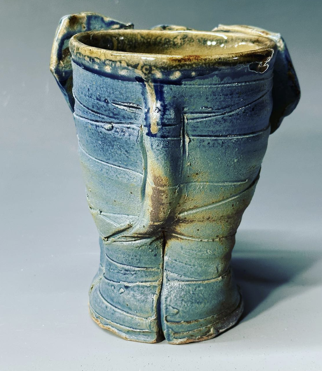 A ceramic elephant mug back view with a flared rim, decorated with uneven blue, green, and beige glaze, and textured with horizontal ridges and cracks.