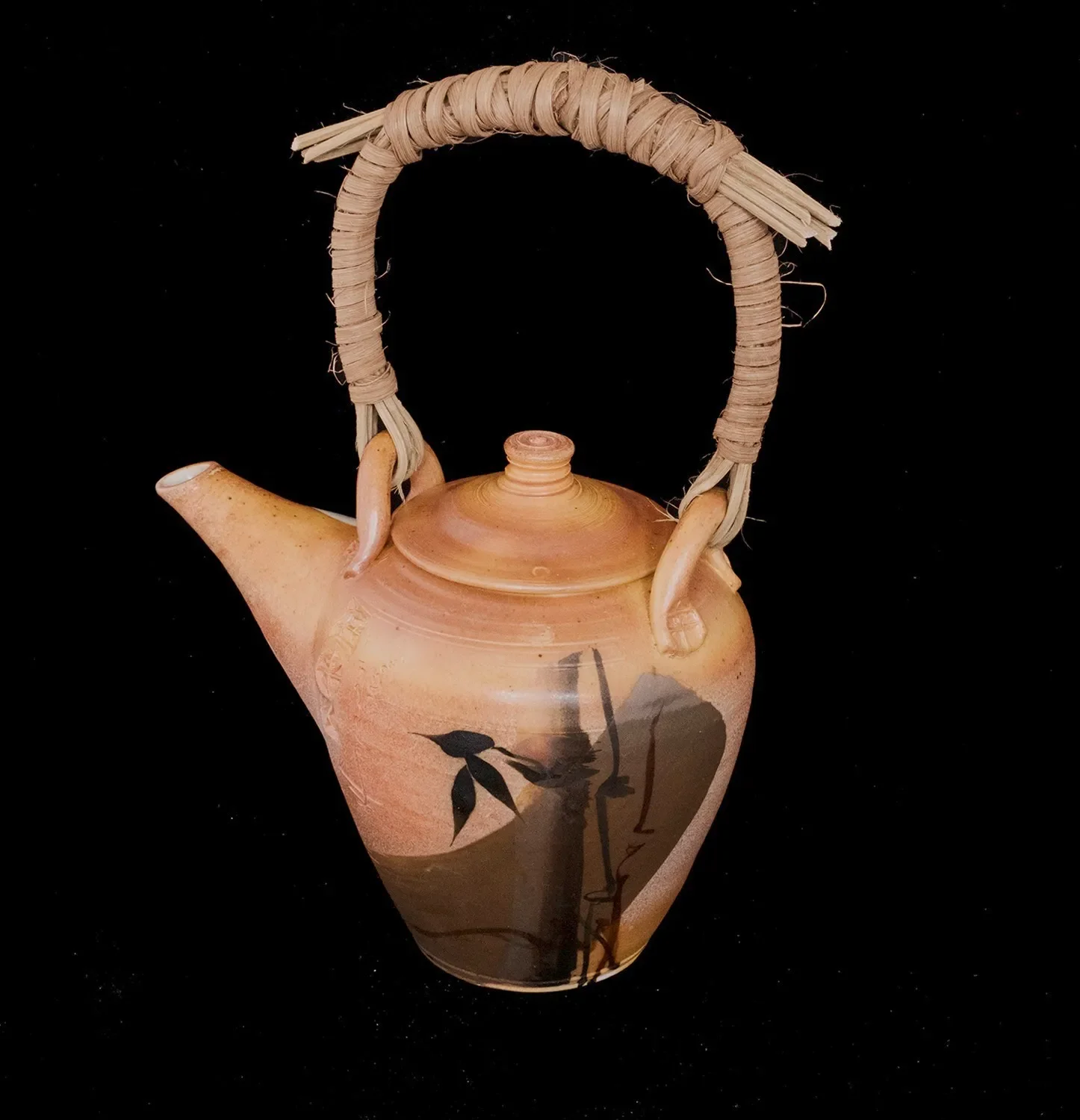 A ceramic teapot with a painted design of black leaves and branches, and a handle made of wrapped straw or bamboo, set against a black background.