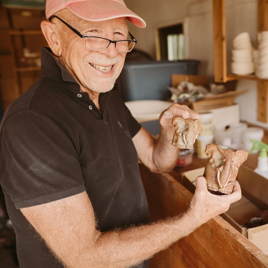 John Hunter smiling and holding two small, carved elephant sculptures in a workshop with shelves of pottery and ceramic supplies.