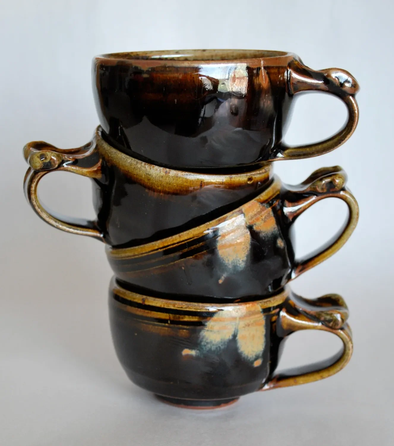 Three stacked ceramic cups with handles, glazed in black and brown with a glossy finish, reflecting light against a plain white background.