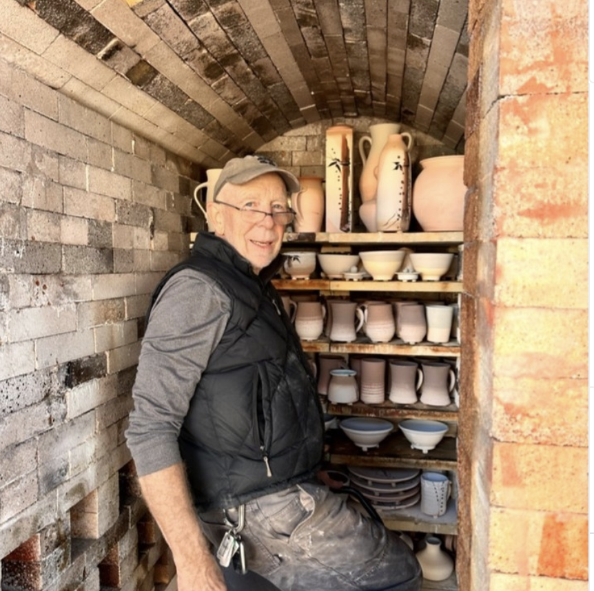 John Hunter wearing a gray cap and black vest, standing inside a brick kiln surrounded by shelves of various pottery, including vases, bowls, and pitchers.