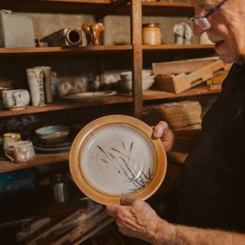 John Hunter holding a ceramic plate, in a room with wooden shelves filled with pottery and ceramics.