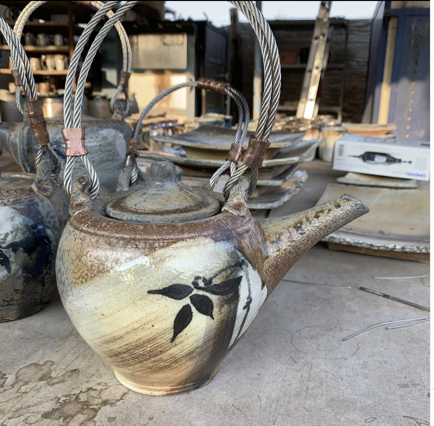 A stoneware ceramic teapot with a sumi-e bamboo design, featuring a steal handle, sitting on a workbench in a pottery studio.