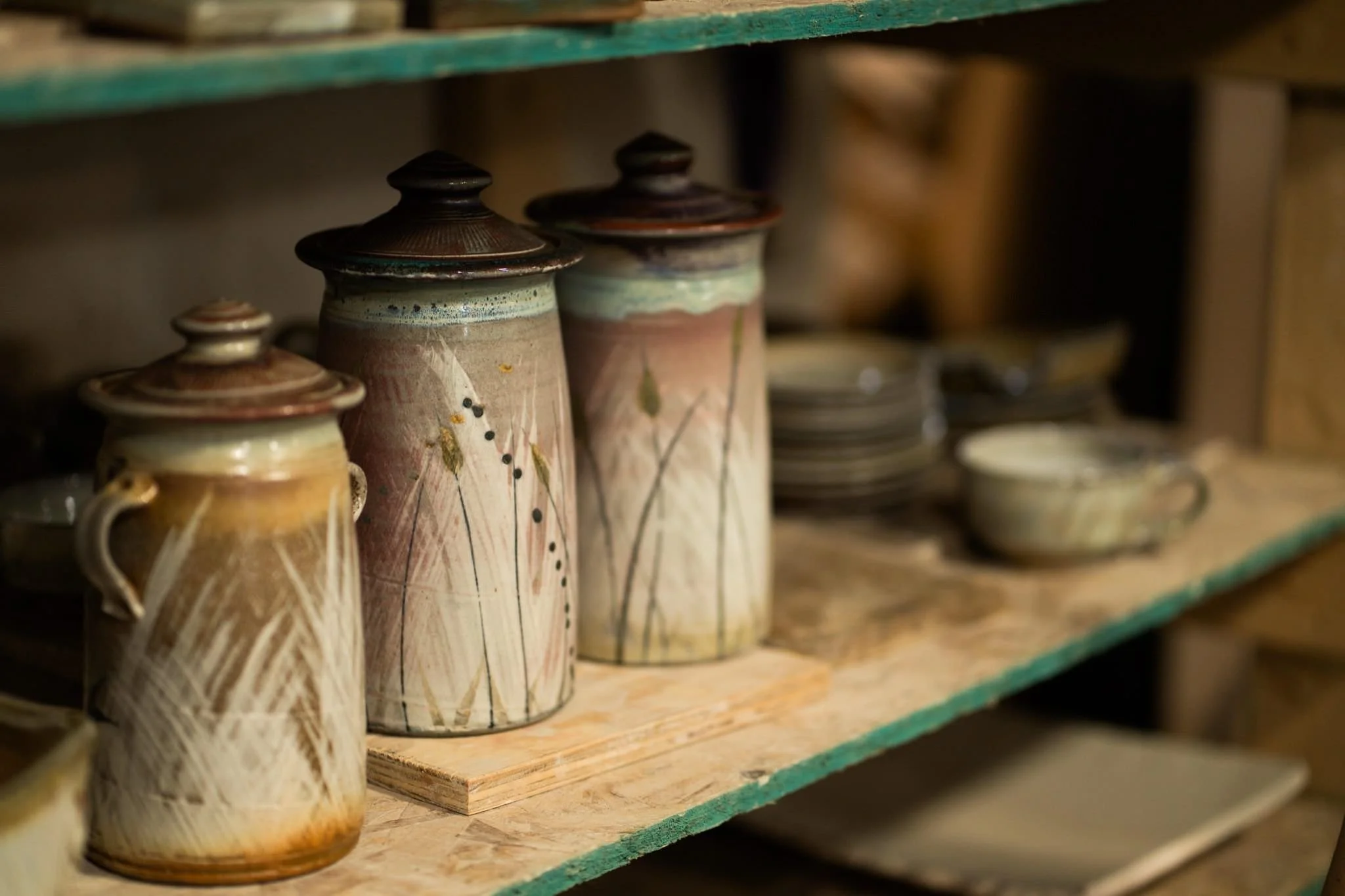 A collection of handcrafted ceramic mugs and containers with lids, displayed on a wooden shelf in a pottery studio or shop.