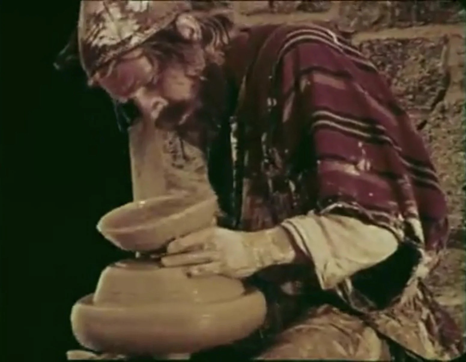 Person wearing a patterned jacket and yellow gloves is using a pottery wheel to shape a clay pot on a sandy ground.