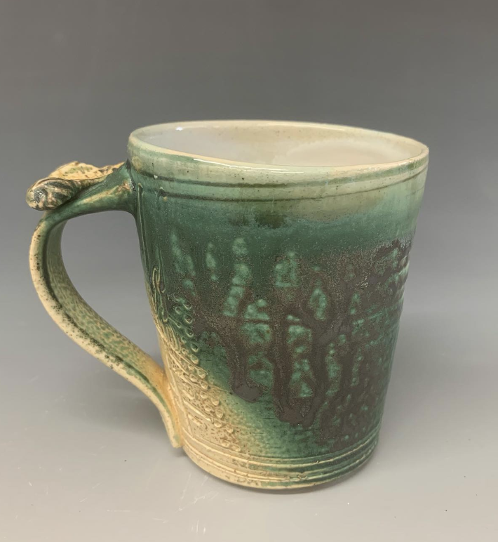 A ceramic mug with a green glaze and textured surface, featuring a small thumb rest on the handle.