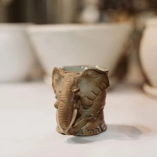 Small elephant-shaped ceramic mug, positioned on a white surface with blurred background.