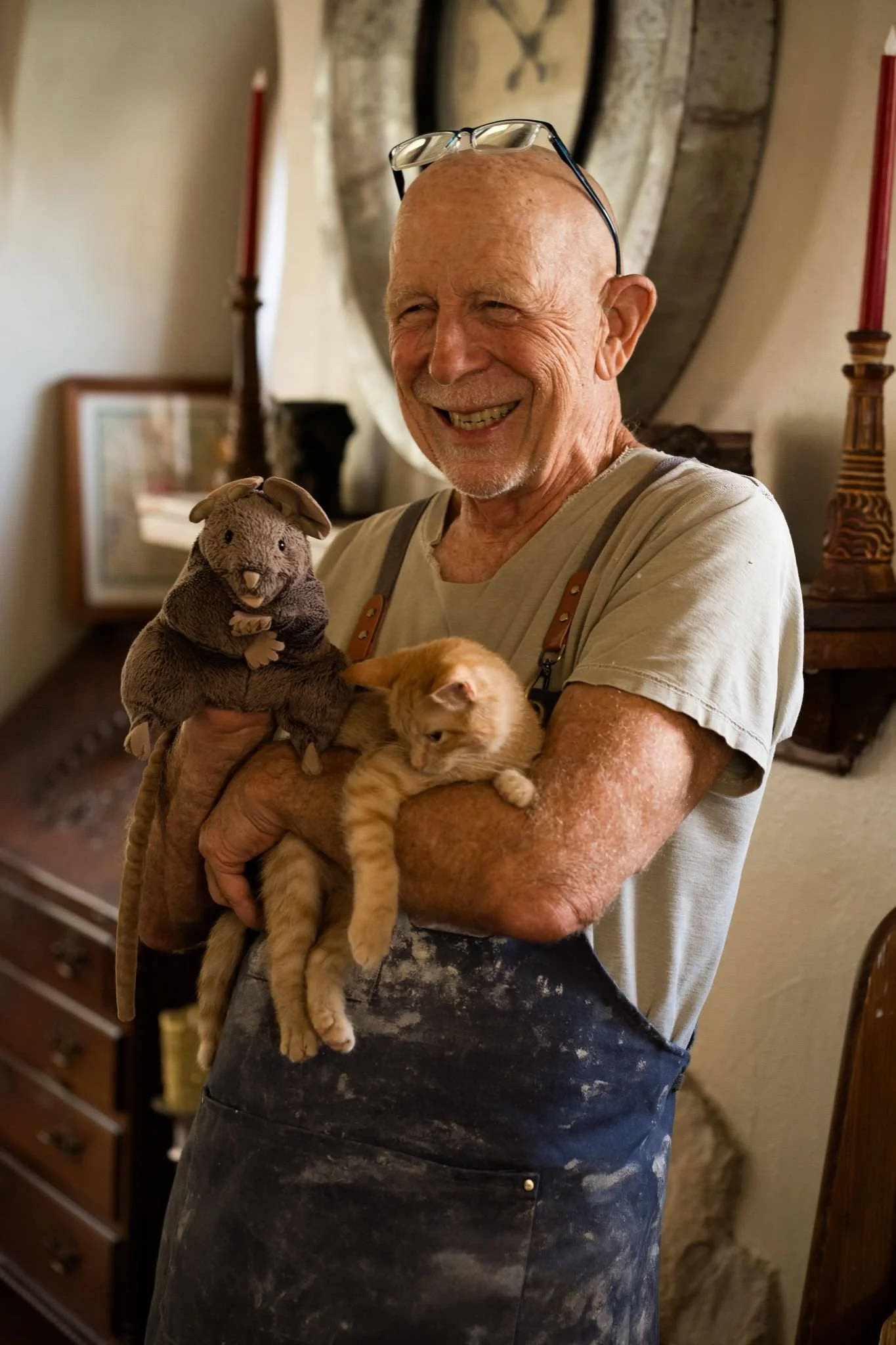 Smiling elderly man holding two kittens, one plush rabbit toy, in a cozy home setting with candles and decorative items in the background.