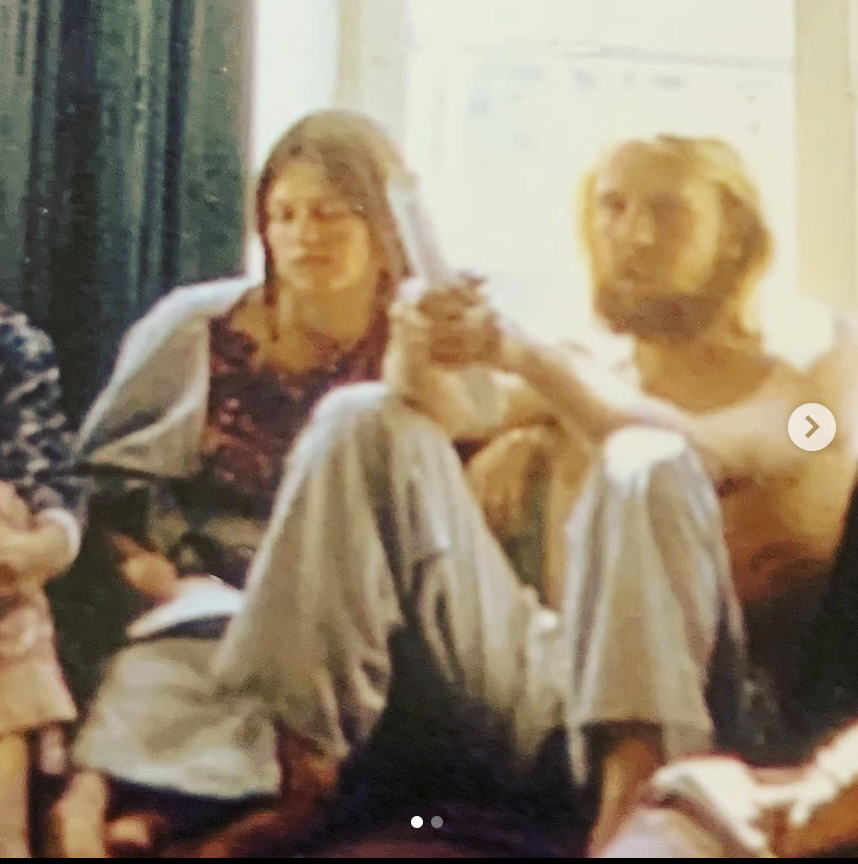 sitting on a couch in a living room. One woman has shoulder-length hair and is wearing a dark patterned shirt, holding a drink. The other person has blonde hair and is wearing a light-colored flannel shirt,