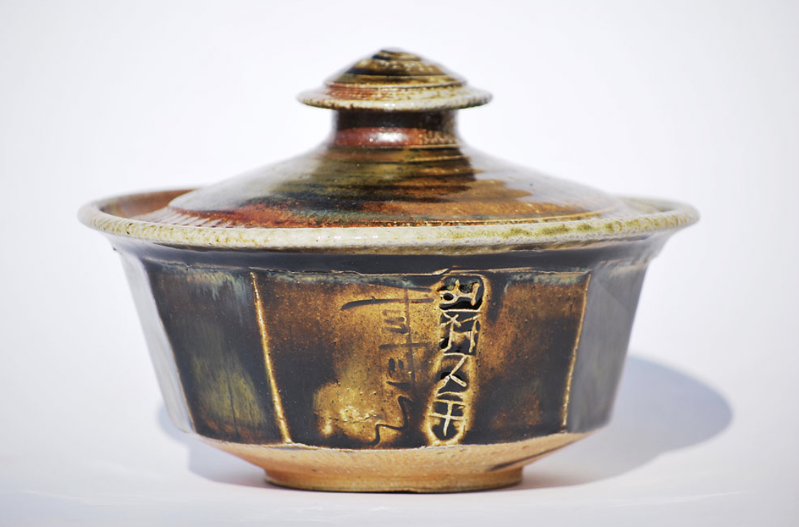 A traditional Japanese ceramic caserol with a lid, featuring Japanese characters and a glazed, earthy-tone finish.