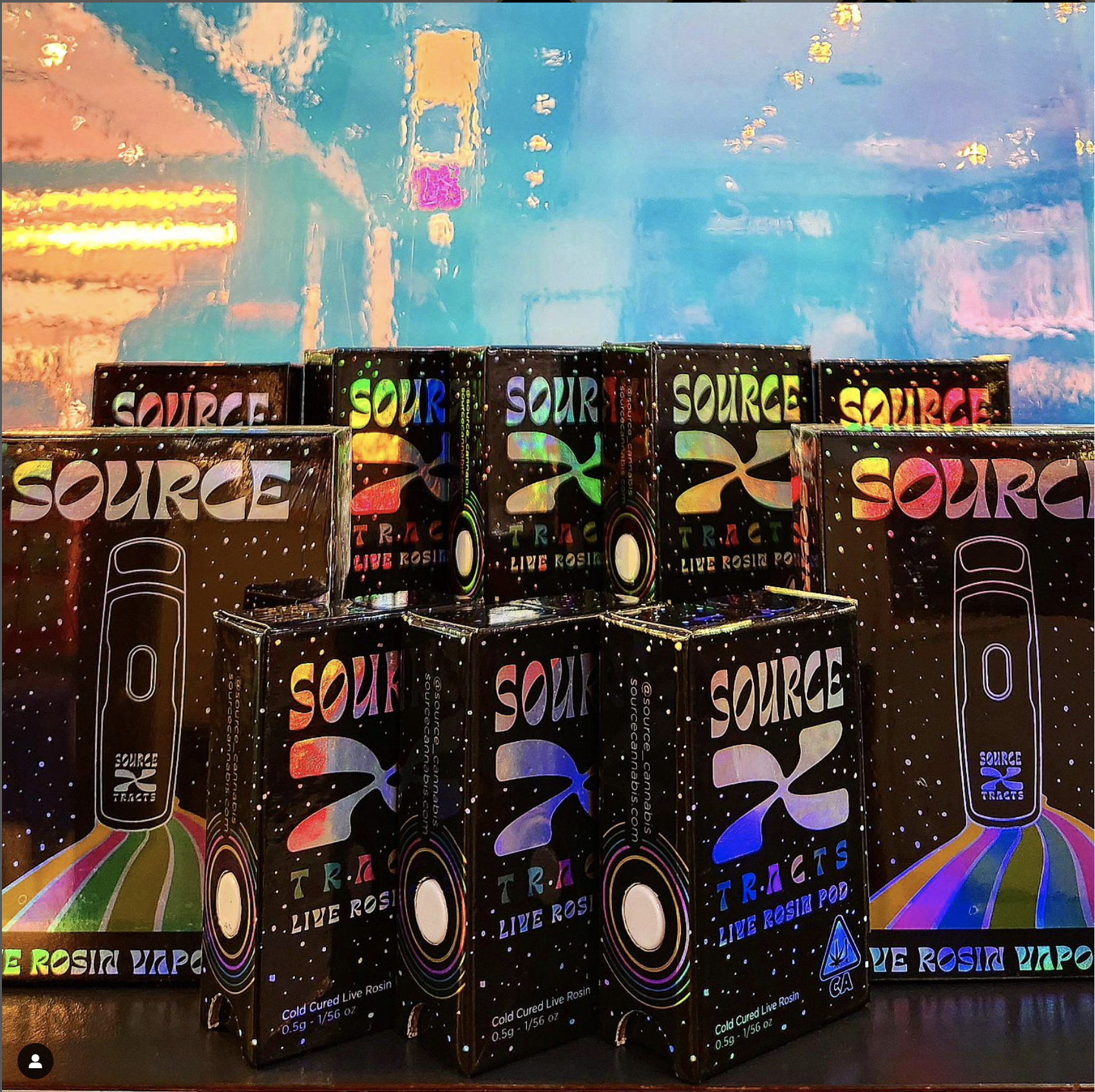 A group of colorful "SOURCE XTRACTS" Live Rosin Vape Pods displayed in front of a bright and abstract background. The packaging features holographic designs with a logo and text detailing the product as "Cold Cured Live Rosin."