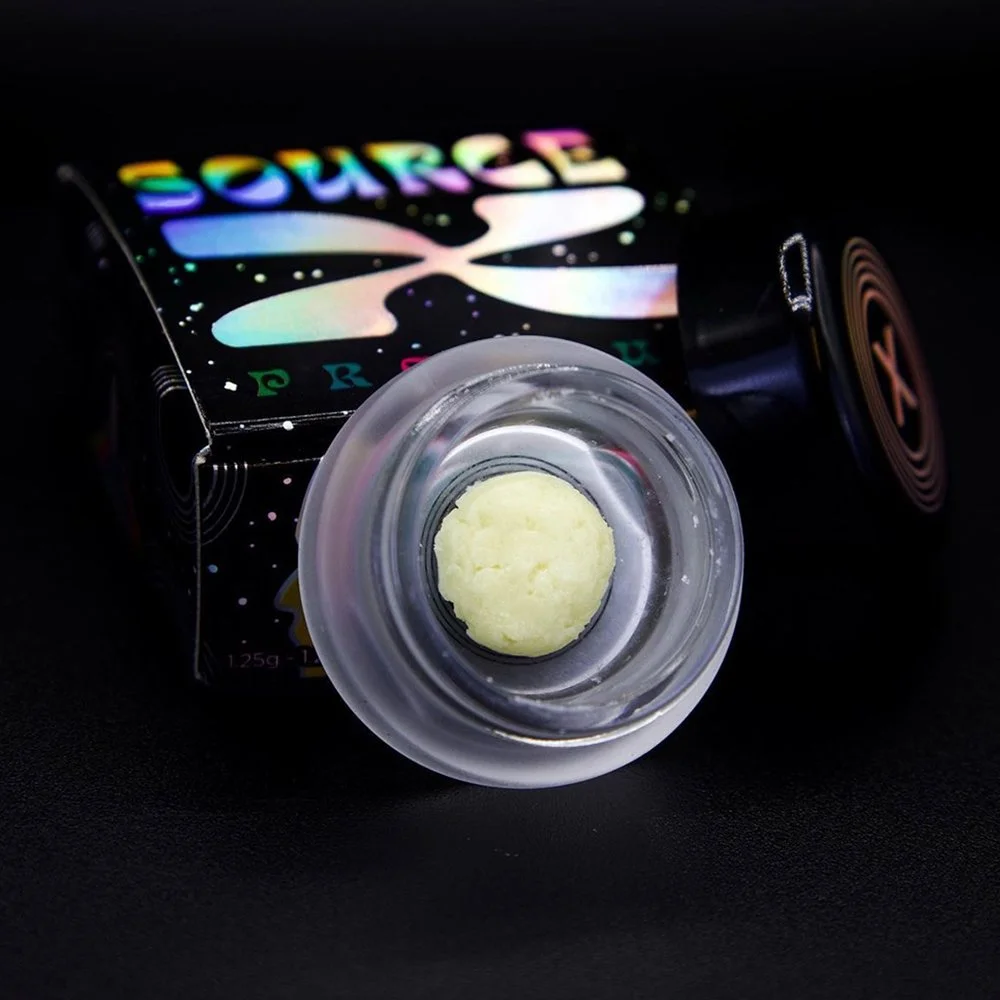 Close-up of a small container holding a round, cream-colored substance, with a colorful, black box labeled 'SOURCE' in the background.