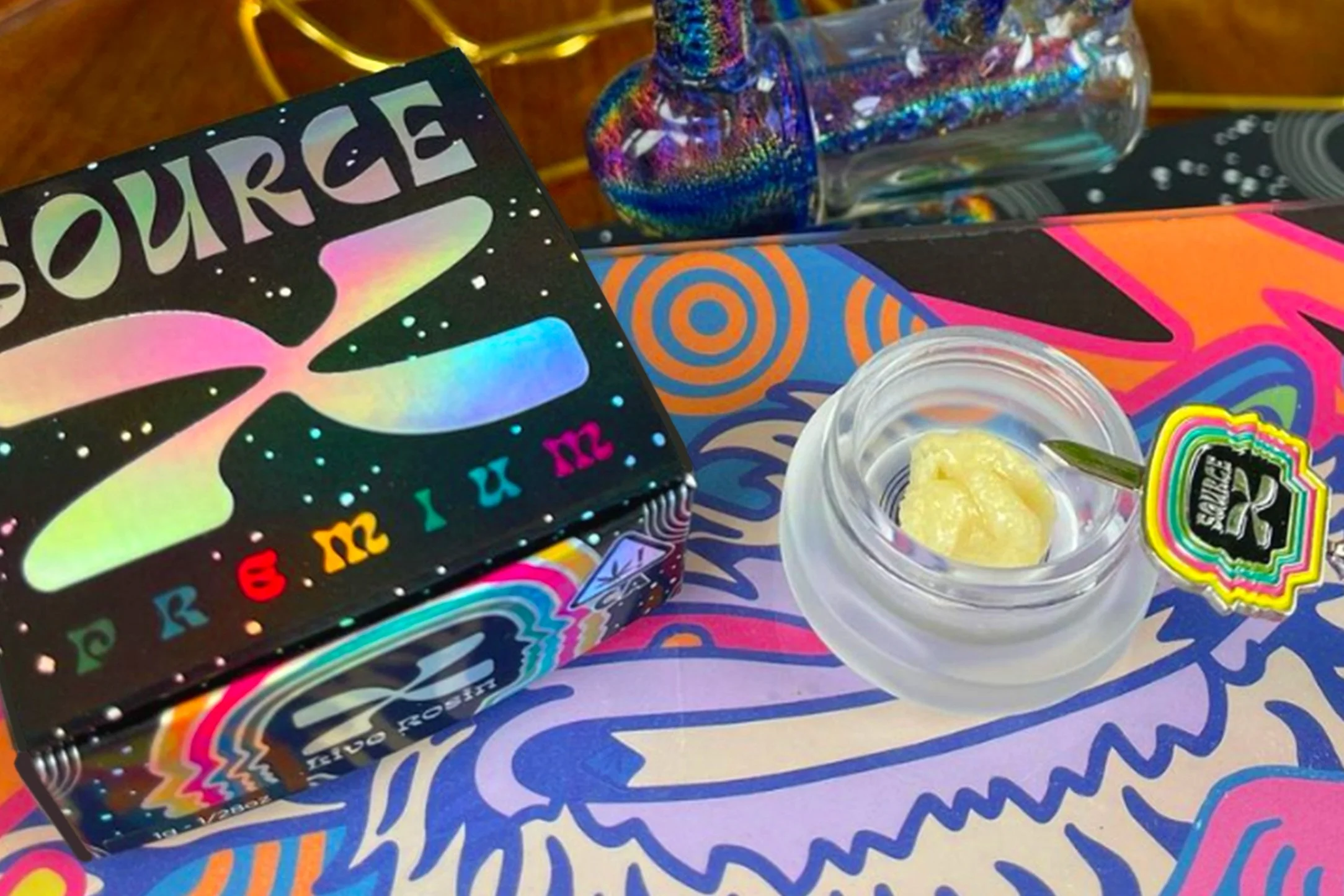 Colorful cannabis product packaging with a glass container of cannabis concentrate and a dab tool on a psychedelic-themed surface.