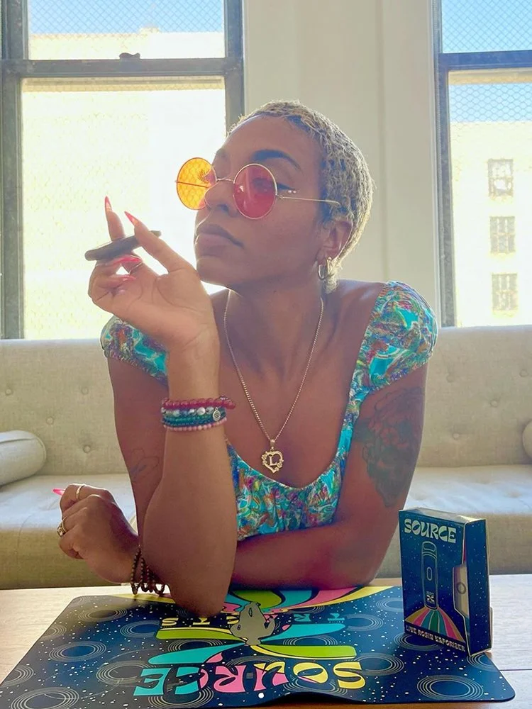 A person with short blond hair wearing red round sunglasses and a colorful top, holding a smoking device. They are leaning on a table with a Source vape box and a psychedelic mat featuring a cartoon cat design. Bright sunlight streams in from a large window behind them.
