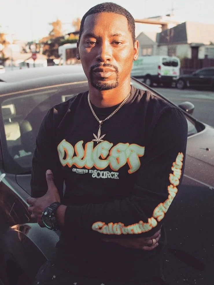 Man standing in front of a car with crossed arms, wearing a black T-shirt with "QUEST" text and a necklace with a marijuana leaf pendant. Background shows a street scene with buildings and parked vehicles.