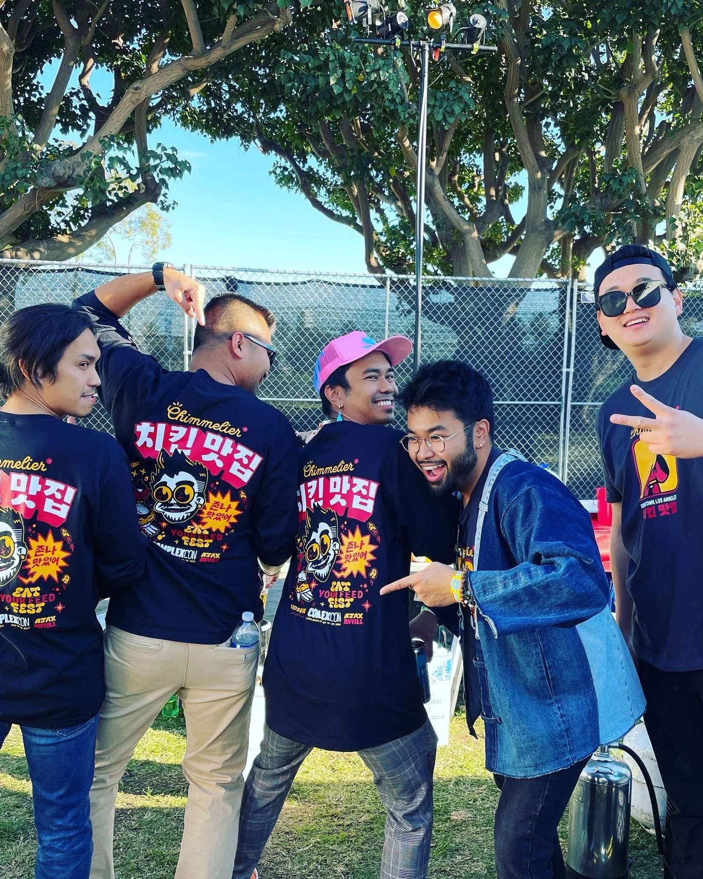 Group of people outdoors, wearing black shirts with colorful graphics and Korean text, making playful poses near a fence and trees.