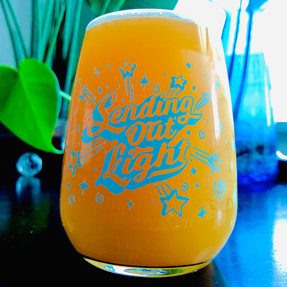 A glass filled with orange juice with "Sending Out Light" written on it in decorative letters, surrounded by star and swirl designs. The background includes a green plant leaf and blue objects, likely vases, under natural light.