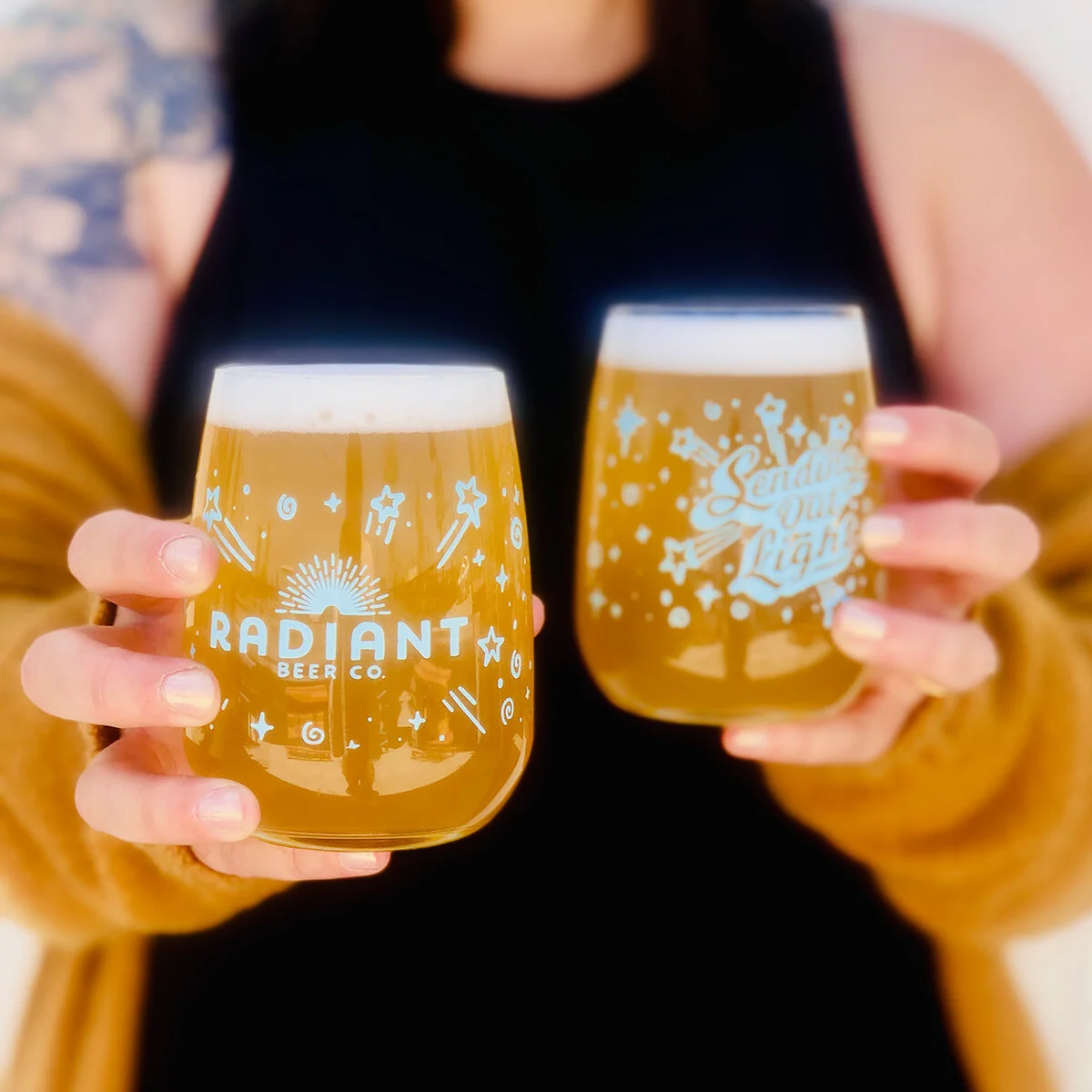 Two hands holding glasses of beer with "Radiant Beer Co." and "Sending Out Light" logos, featuring star and swirl designs. Person wearing black top and mustard-colored cardigan.