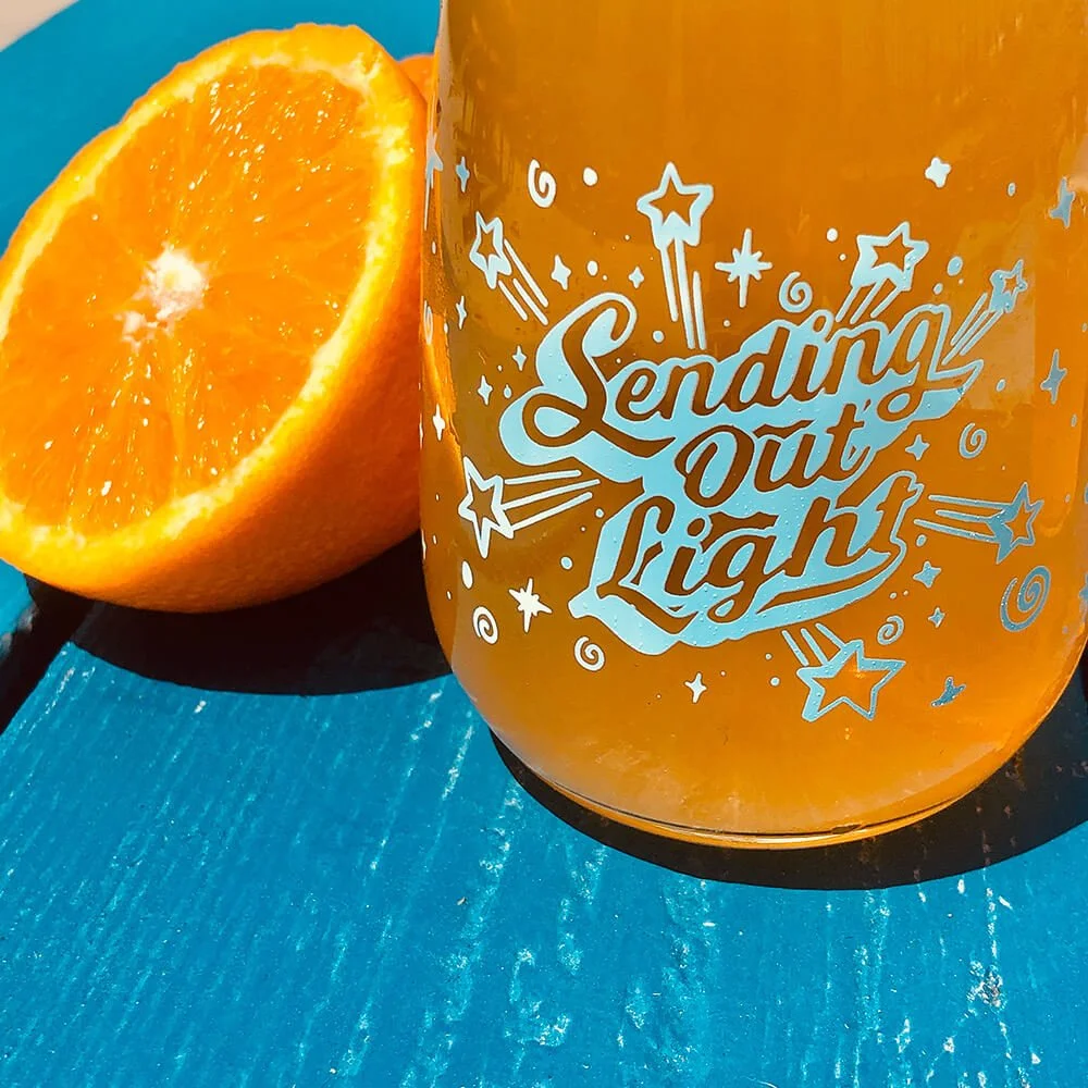 Orange drink with "Sending Out Light" text in blue on glass, next to half orange, on blue surface.