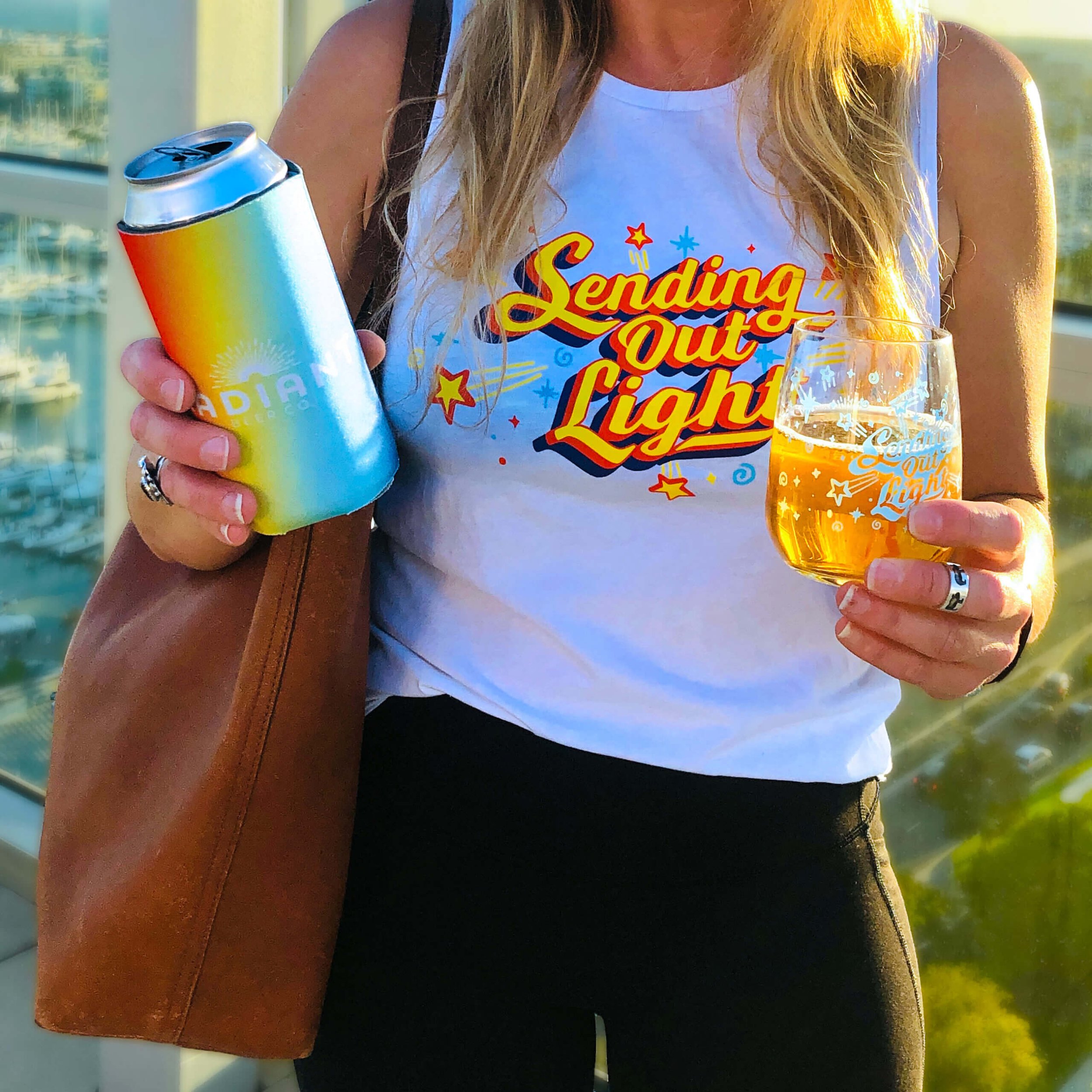 Person holding a drink in a glass, wearing a "Sending Out Light" tank top, and holding a rainbow-colored can cooler.