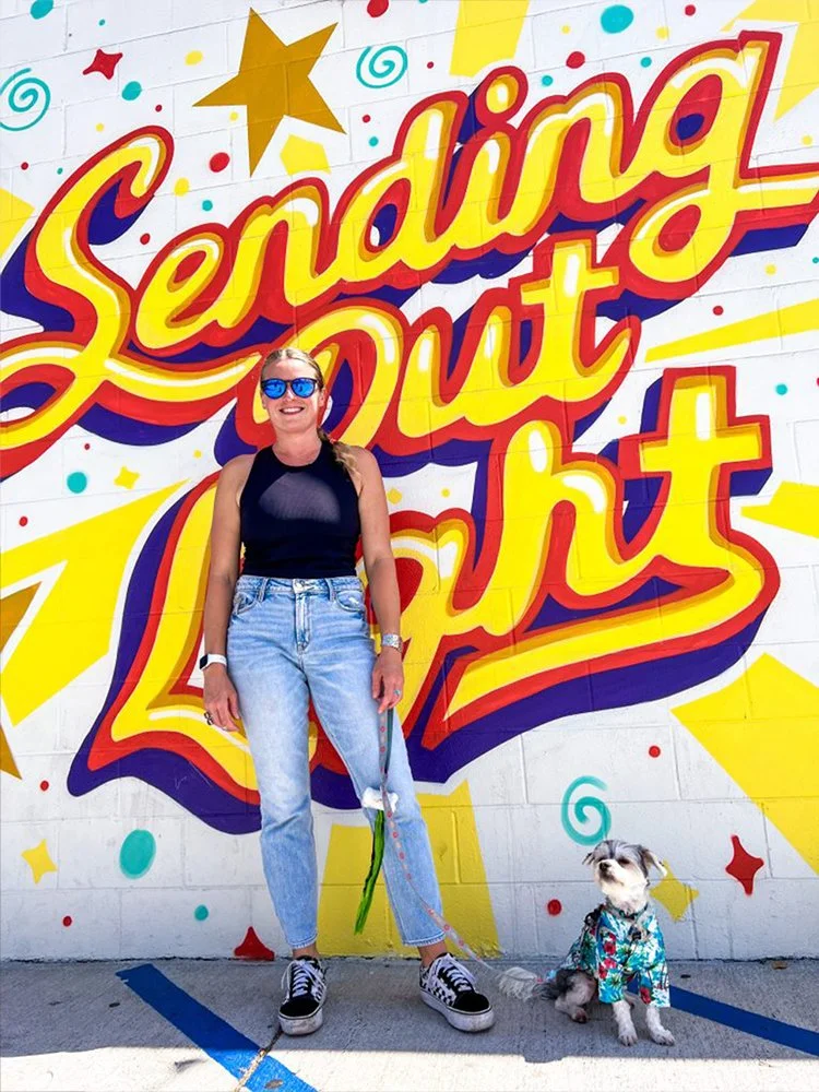 Woman in sunglasses standing next to a small dog in a colorful shirt, in front of a mural with the text 'Sending Out Light' and colorful stars and swirls.