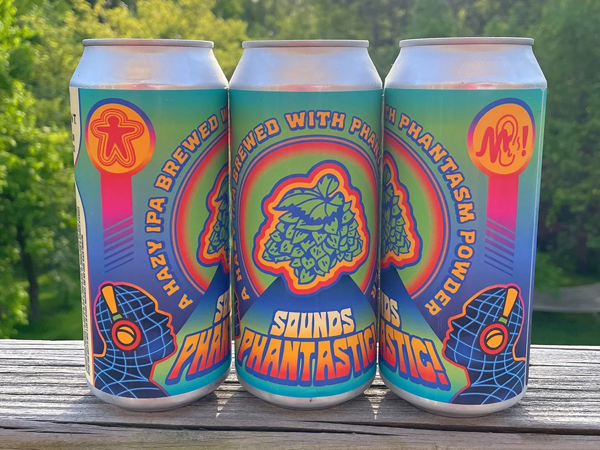 Three cans of Hazy IPA with colorful abstract artwork, featuring hop illustrations and graphic designs on the label, standing on a wooden surface with a green outdoor background.