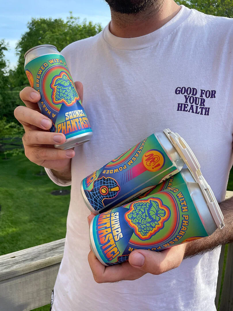 Person holding cans of "Sounds Phantastic" beverage with colorful design, wearing a white shirt with "Good For Your Health" printed on it.