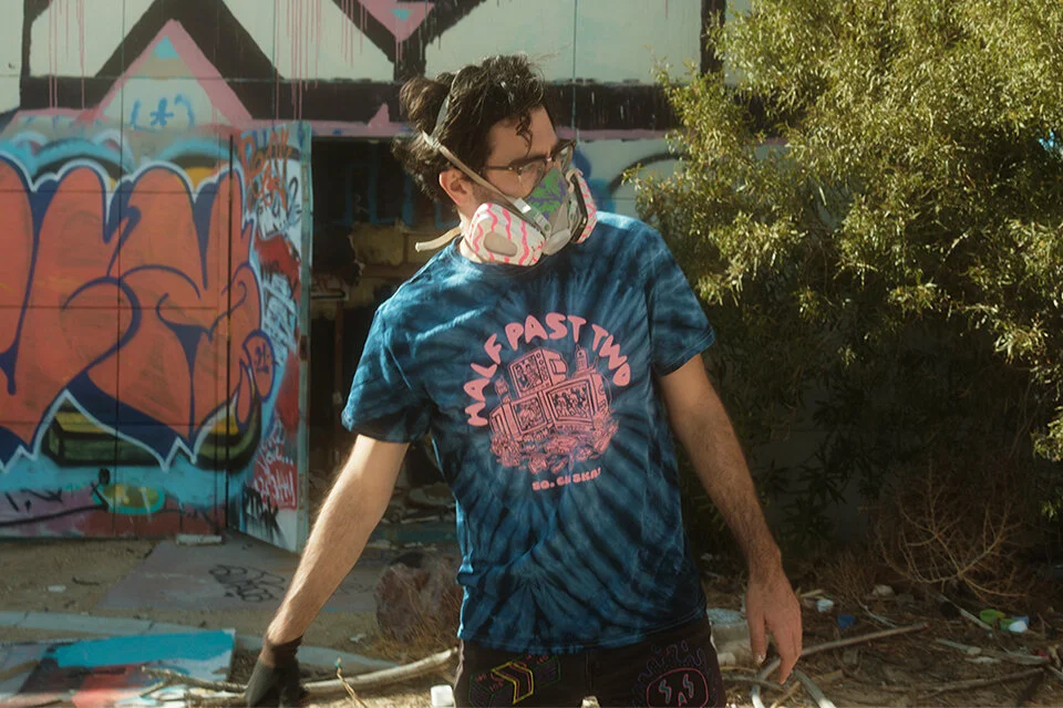 Person wearing a tie-dye shirt and a respirator mask standing near graffiti-covered wall.
