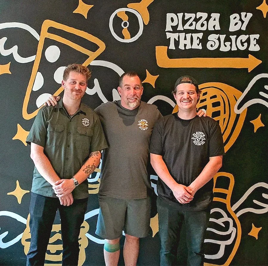 Three men posing in front of a mural with pizza-themed art at a pizzeria.