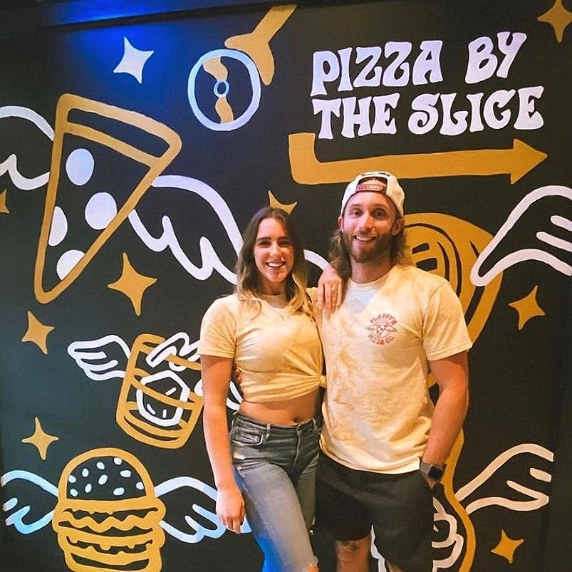 Two people standing in front of a wall with pizza-themed art, featuring phrases like "Pizza by the Slice" and illustrations of pizza slices and other food items with wings.