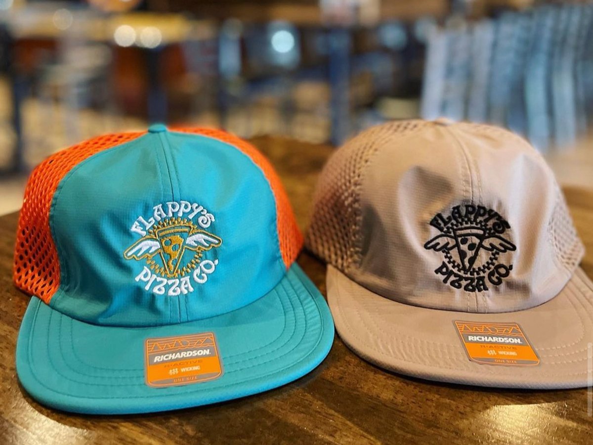 Two trucker hats on a wooden table, one teal with a red mesh back and the other beige, featuring "Flappy's Pizza Co." logo on the front. Both have "Richardson" brand stickers on the brims.
