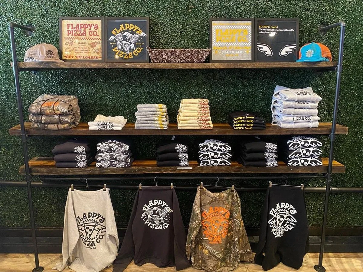 Display of Flappy's Pizza Co. merchandise including t-shirts, hoodies, hats, and framed signs on wooden shelves against a green backdrop.