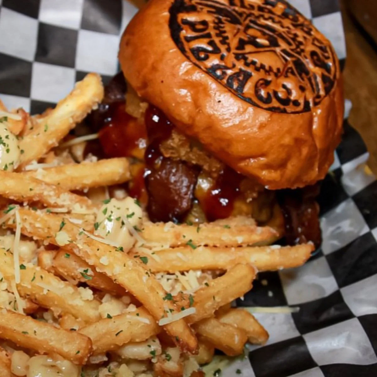 Gourmet burger with branded bun and seasoned French fries on checkered paper.