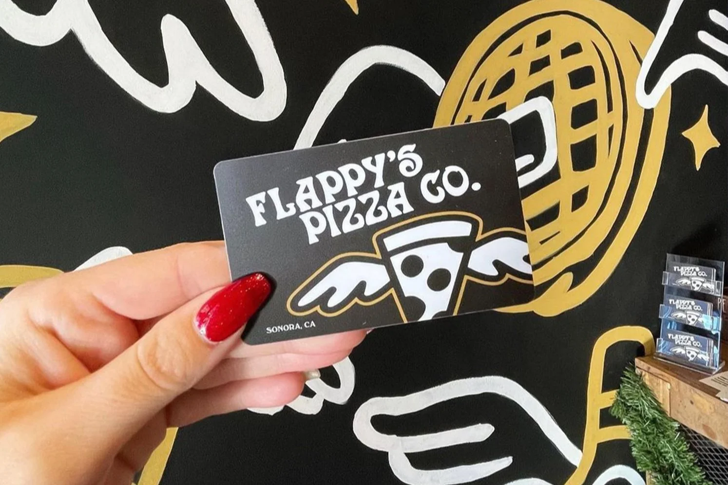 Hand holding a Flappy's Pizza Co. gift card with a background mural of a winged pizza slice and golden design.