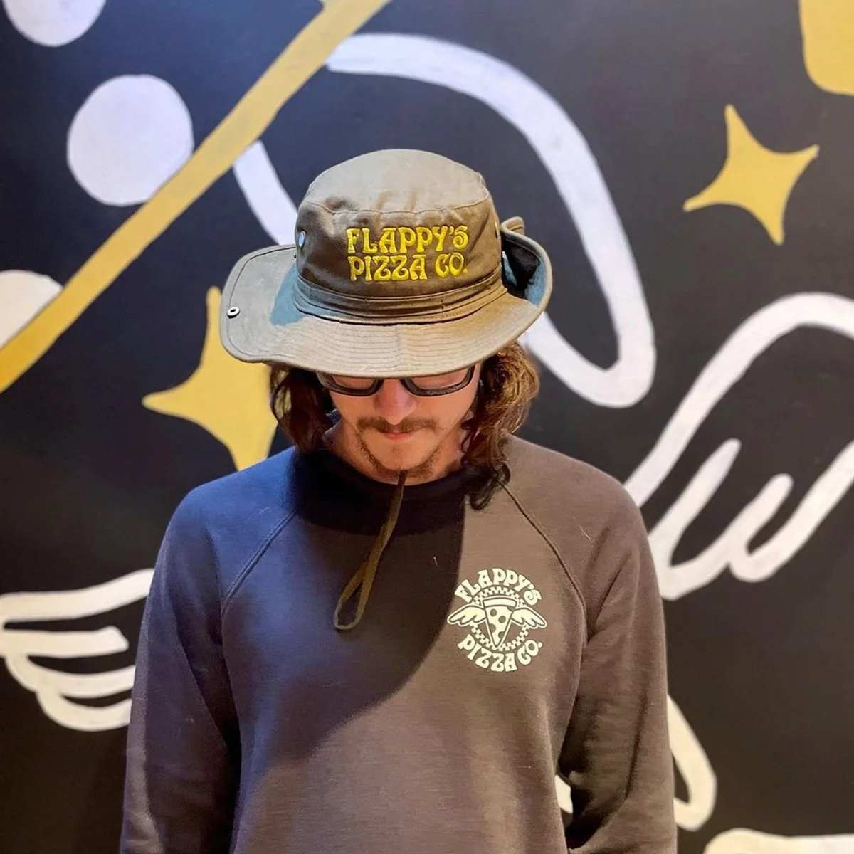 Person wearing a Flappy's Pizza Co. hat and sweatshirt, standing in front of a wall with artistic black, yellow, and white designs.