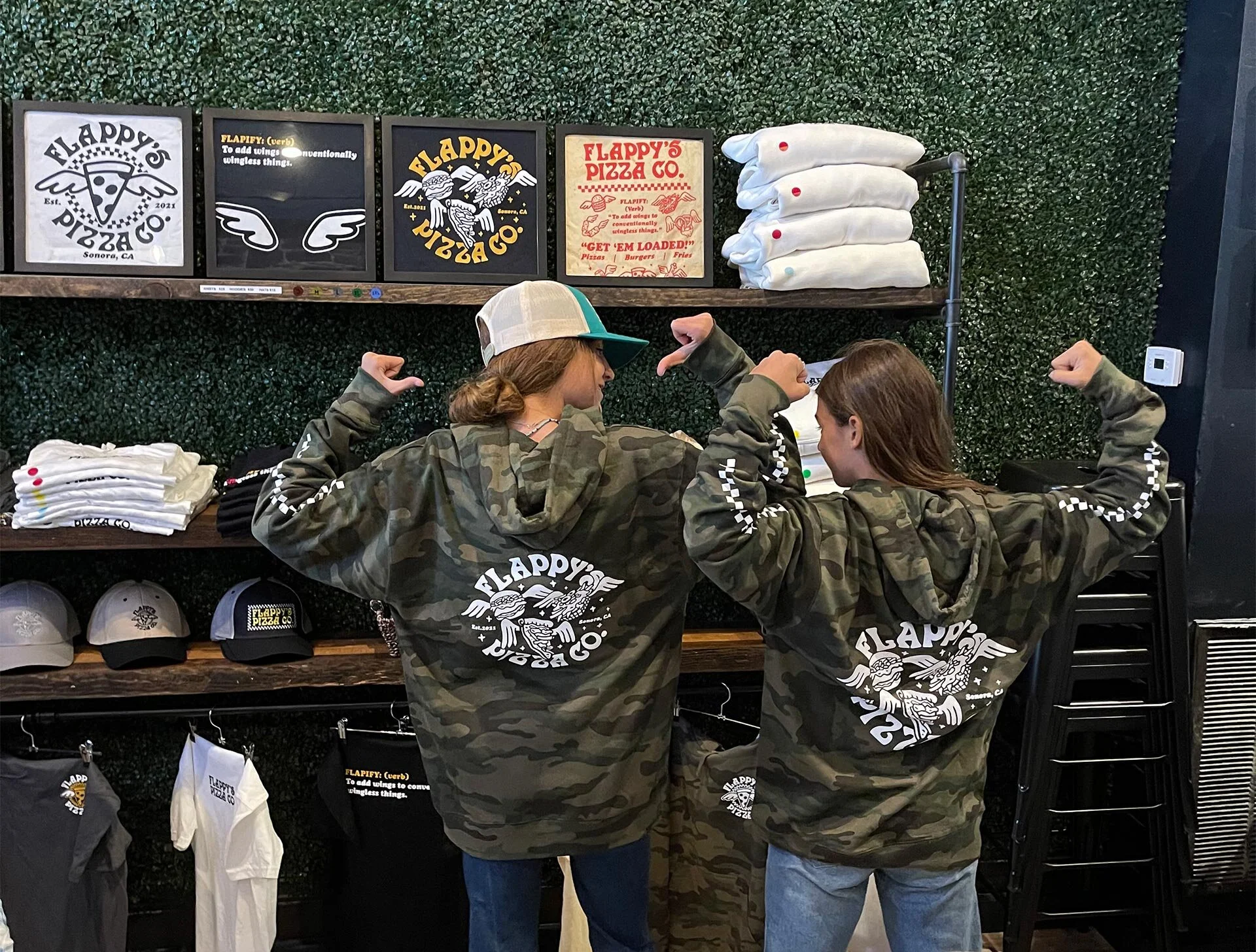 Two people wearing hooded camo jackets with "Flappy's Pizza Co." logo, flexing their arms, stand in front of merchandise shelves displaying hats, shirts, and framed artwork at a pizza store.