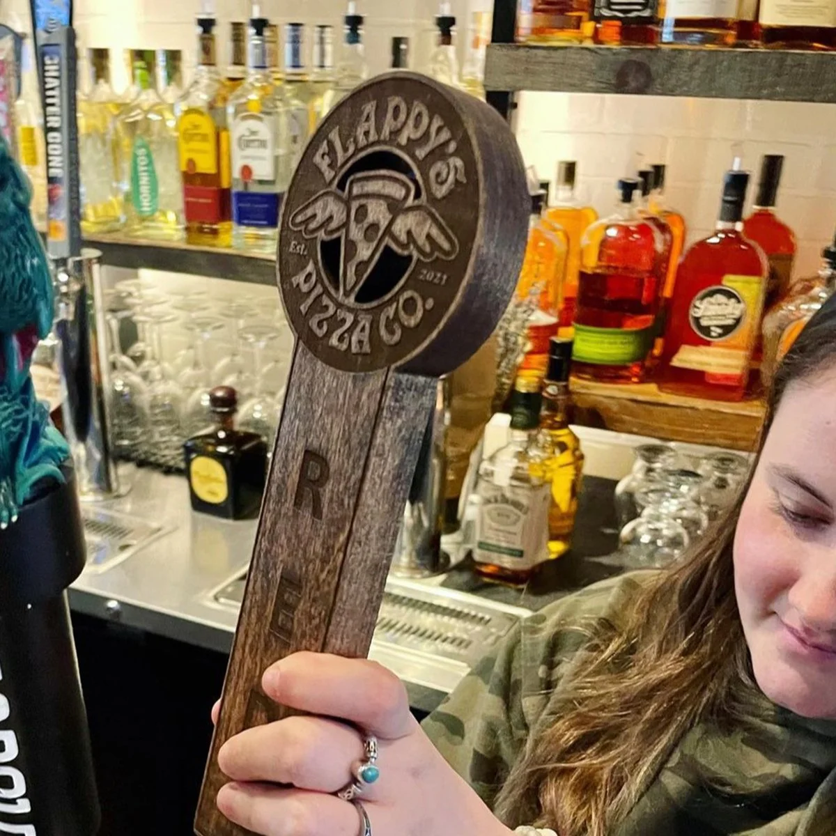 A person holding a beer tap handle labeled "Flappy's Pizza Co" in a bar setting with bottles of liquor in the background.