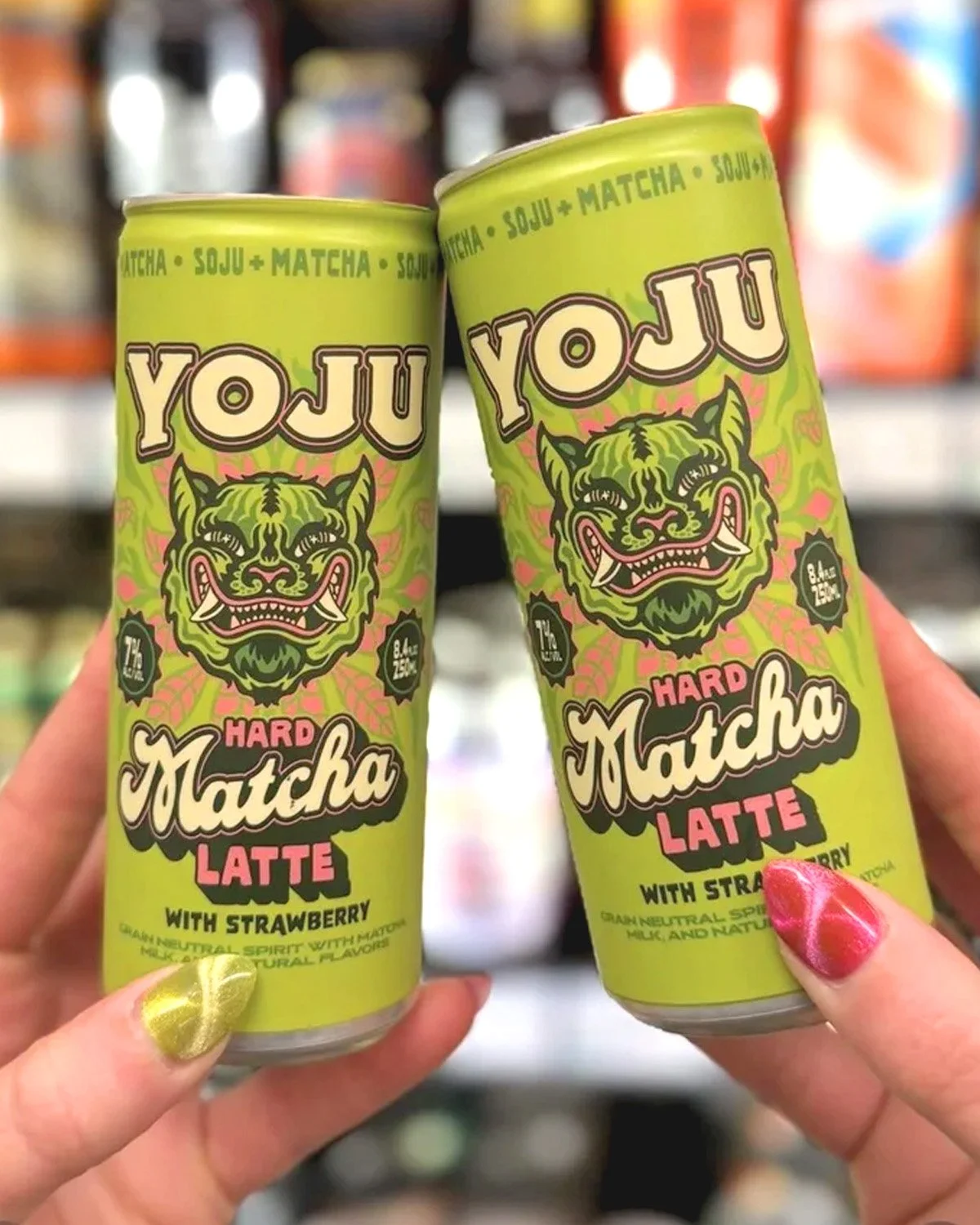 Two Yoju Hard Matcha Latte cans held in front of a retail cooler, showing the green tiger packaging design.