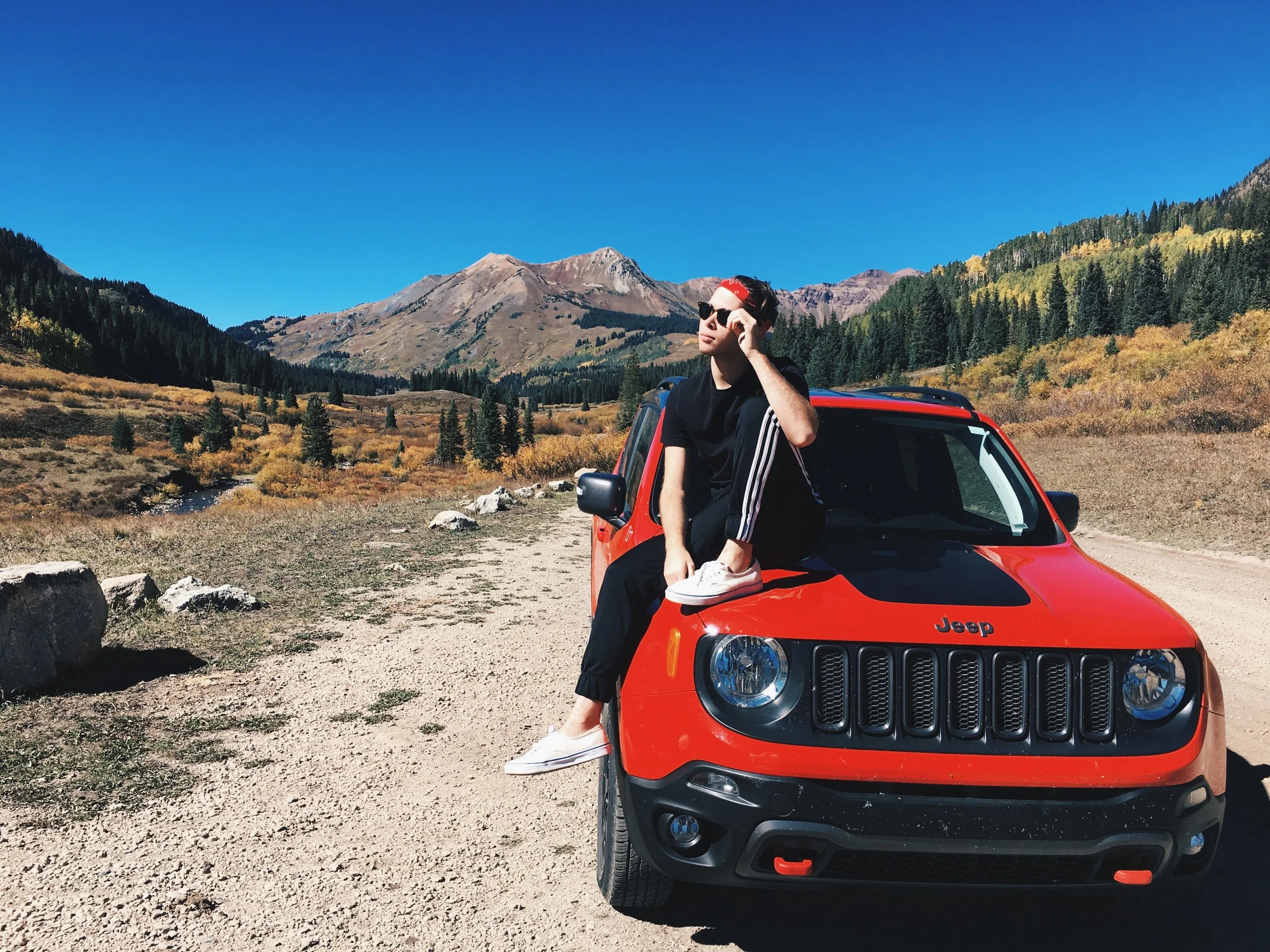 A young person sitting on the front of a red Jeep parked in a mountainous landscape with clear blue sky, trees, and mountains in the background.