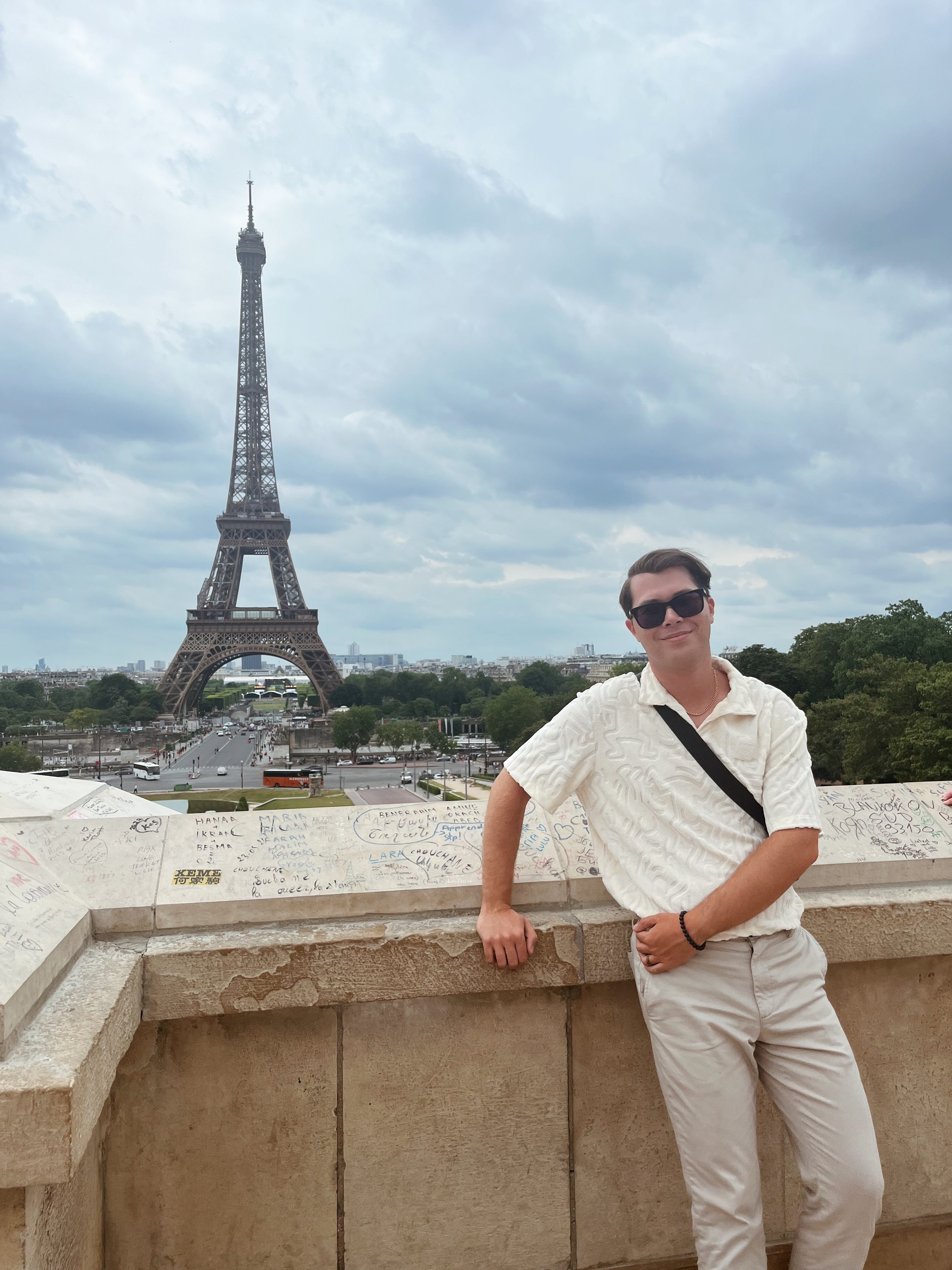 A person wearing sunglasses and a white shirt with a black sash, standing with one arm resting on a stone ledge, in front of the Eiffel Tower in Paris, France, with cloudy skies overhead and a cityscape and greenery in the background.