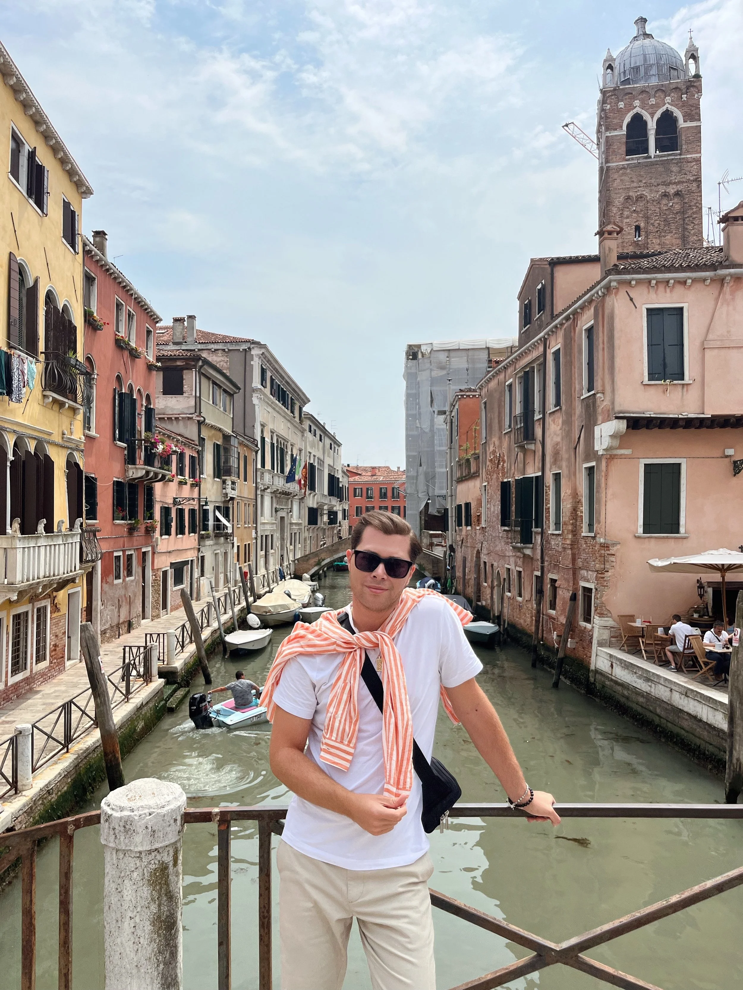 A young man wearing sunglasses, a white t-shirt, khaki pants, and a light orange striped shirt tied around his shoulders standing on a bridge over a canal in Venice, Italy, with colorful buildings and boats in the background.