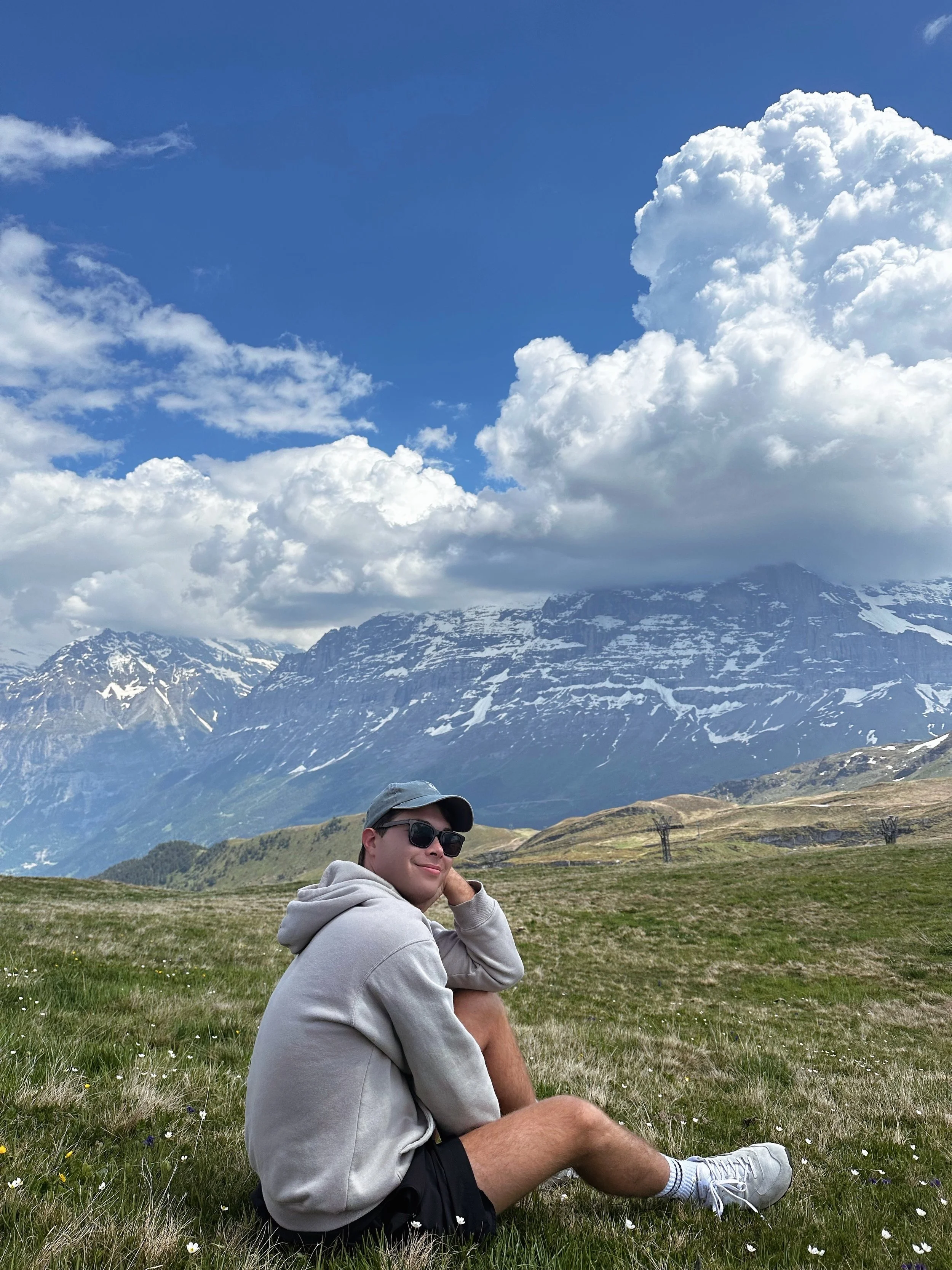 A young man sitting on a grassy meadow with wildflowers, wearing a hoodie, shorts, sneakers, sunglasses, and a cap, with snow-capped mountains and clouds in the background.