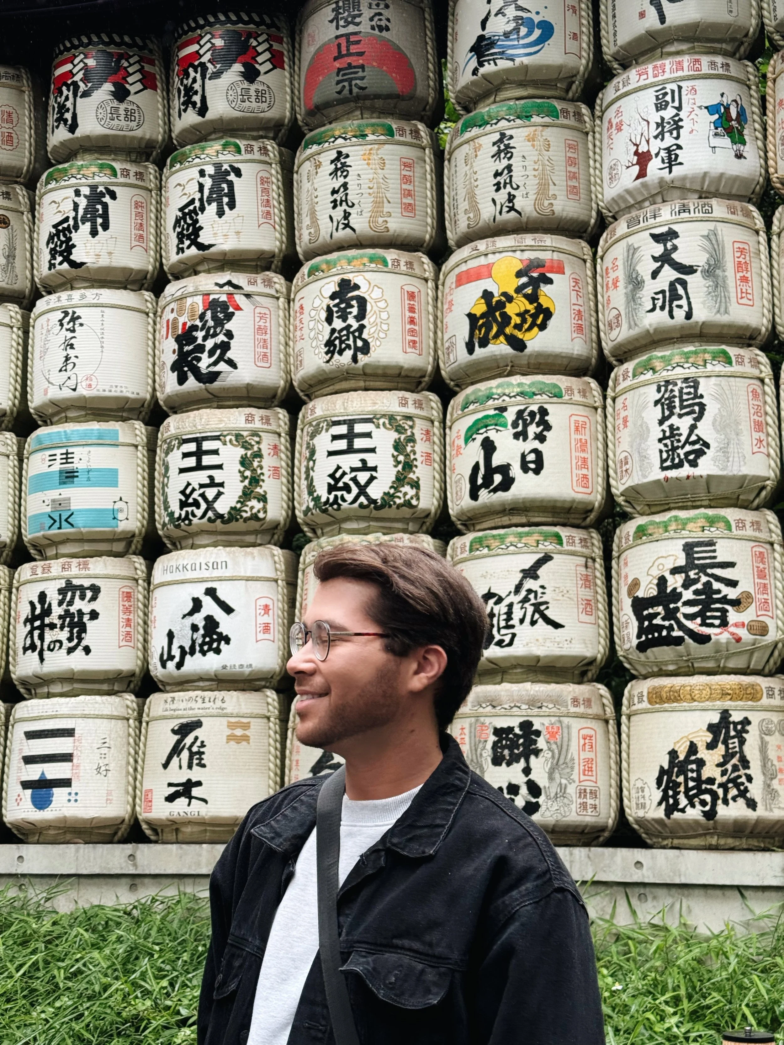 A man with glasses and dark hair smiling, wearing a black jacket and a white shirt, standing in front of a wall of traditional Japanese sake barrels, with green grass at the bottom of the image.