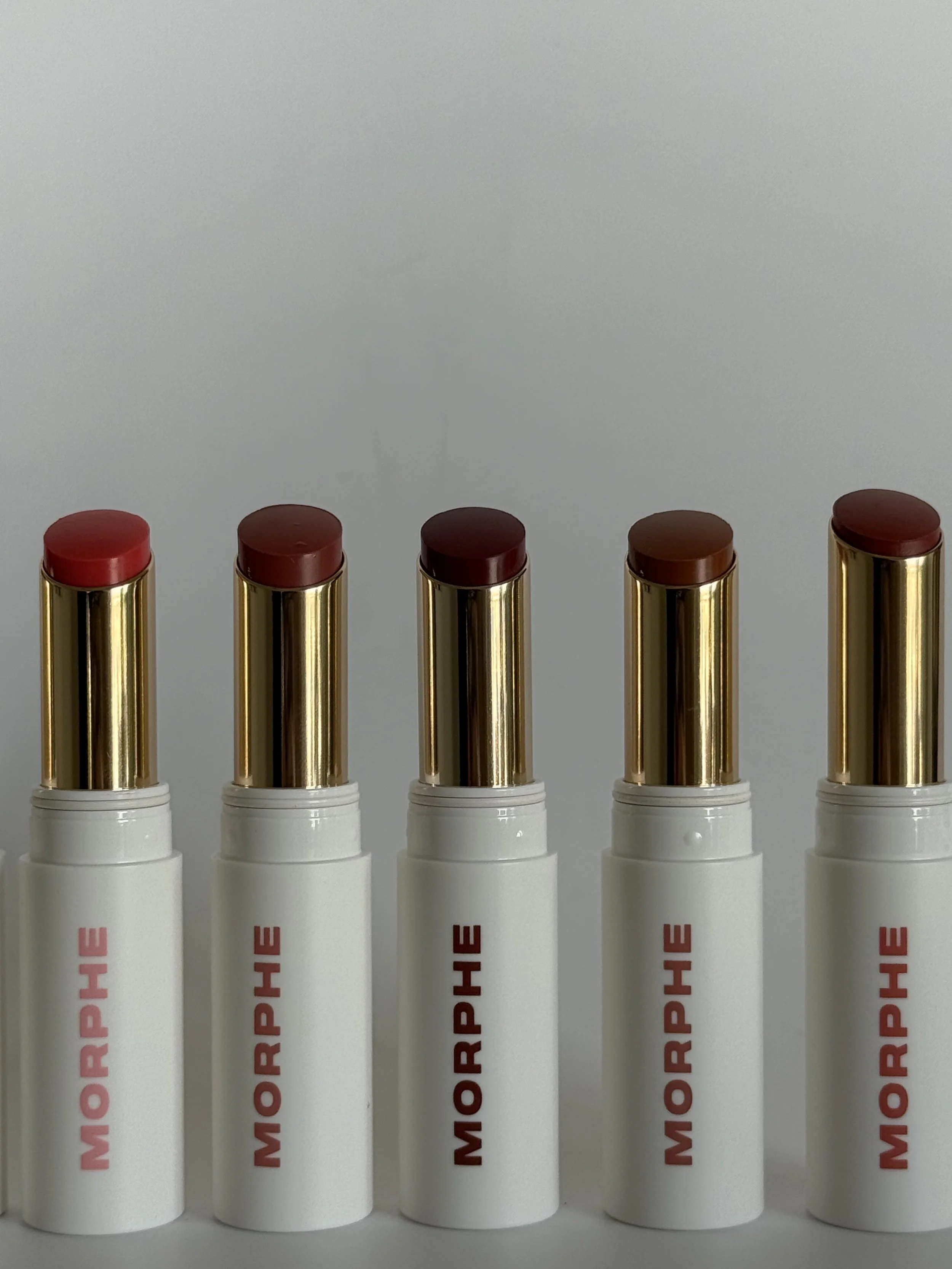 Five white lip balm tubes with gold caps, each labeled 'MOOREHELLO' in pink letters, standing upright against a plain background.